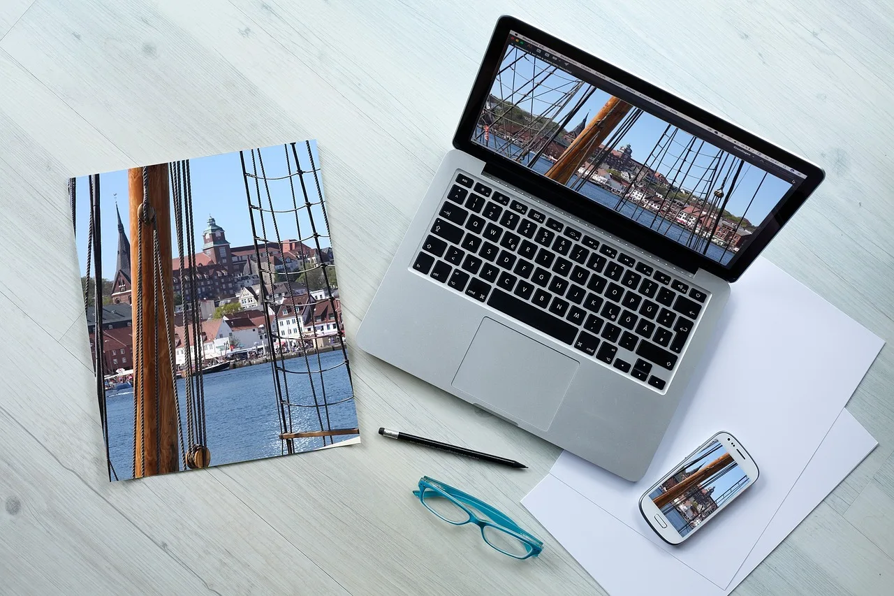 Aufgeklappter Laptop auf einem hellen Tisch, daneben eine Broschüre, ein Smartphone, eine Brille und ein Stift