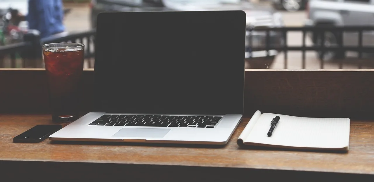 Aufgeklappter Laptop und ein Notizbuch mit Stift auf einem Holztisch, daneben ein Glas